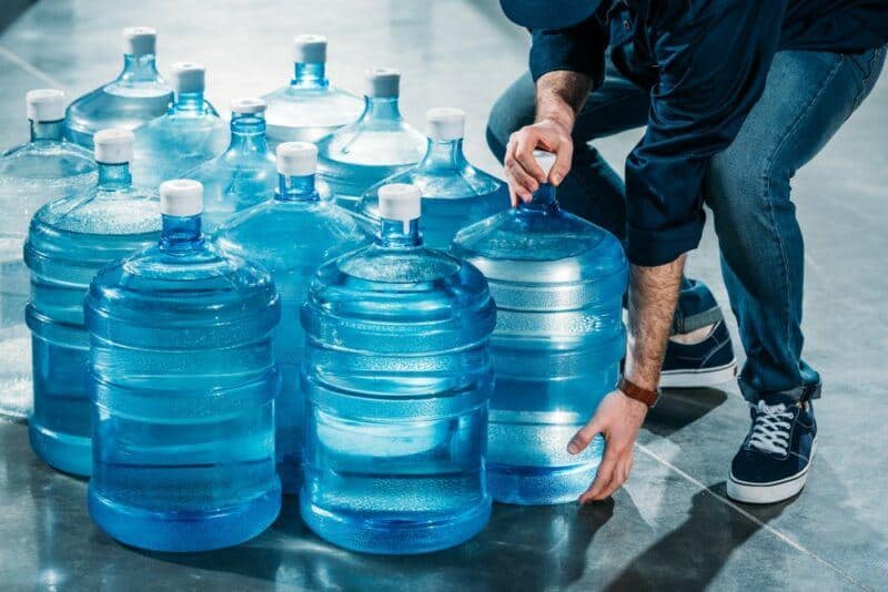 A Collection Of Gallon Water Jugs With A Man Trying To Lift One 800x534